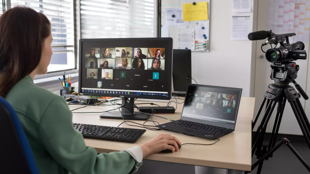 Image of a person sitting at a desk, screen and laptop in front. The screen shows a video conference with multiple other persons participating.